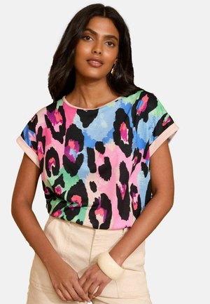 Frau mit dunklem Haar, die ein bunten, abstrakten Tierprint-Shirt mit kurzen Ärmeln und hellbeigen Hosen trägt, steht vor einem schlichten Hintergrund.