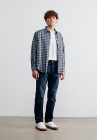 Νεαρός άνδρας που στέκεται, φορώντας γκρι πουκάμισο με καρό πάνω από λευκό μπλουζάκι, σκούρο τζιν και λευκά αθλητικά παπούτσια με καφέ σόλες.