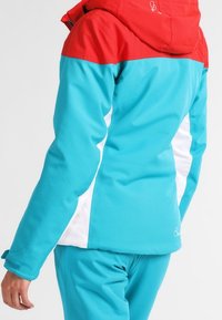 Türkis und rote Skijacke mit Kapuze, ausgestattet mit weißen Seitenpaneelen und verstellbaren Bündchen. Glatte Textur, sportliches Design und lebendige Farben.