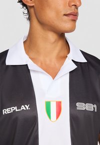 Sort og hvid polo t-shirt med krave, der har et trekantet italiensk flagmærke og "REPLAY" logo på venstre side.