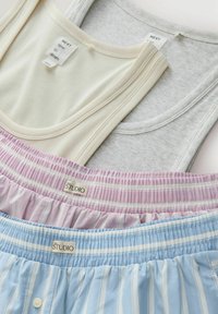 Deux débardeurs à côtes en crème et gris clair avec étiquettes, et deux shorts élastiques à taille rayée en rose et bleu étiquetés "STUDIO".