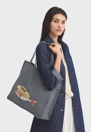 Mujer con abrigo de mezclilla lleva un gran bolso estilo tote a rayas con un gráfico de un pez que lleva un sombrero de paja en la cabeza.