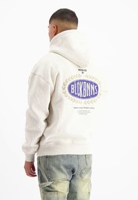 Grijze hoodie met een grote afbeelding op de achterkant, met een blauw ovaal logo en een laurierontwerp. Ribgebreide manchetten en tailleband. Draagt met cargobroeken.