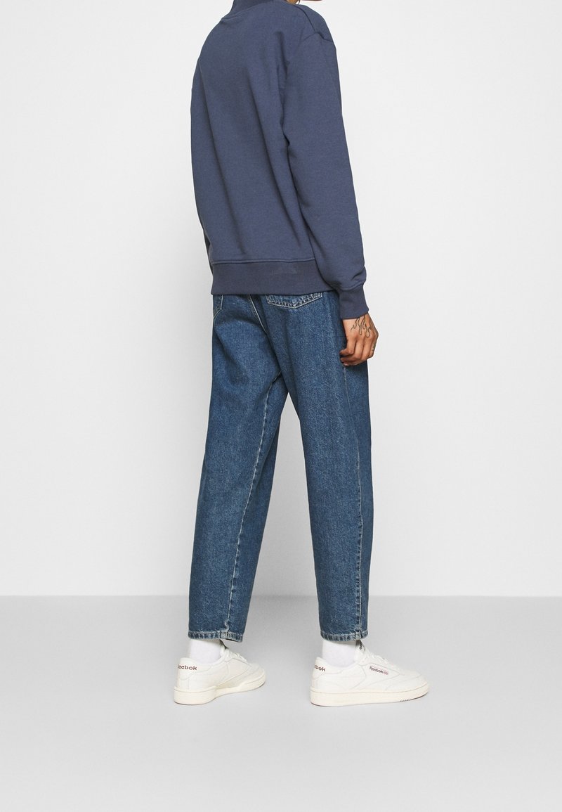 Personne portant un sweat-shirt bleu marine, un jean droit bleu, des chaussettes blanches et des baskets blanches Reebok, debout devant un fond blanc uni.