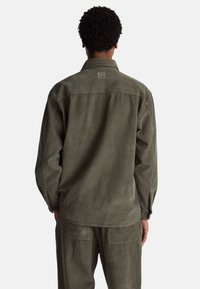 Camicia in velluto a coste verde oliva con maniche lunghe, dotata di colletto a punta e una toppa quadrata con logo sulla parte posteriore del giogo.