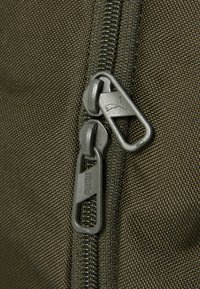 Olijfgroene stoffen tas met twee metalen ritsen, voorzien van Puma-branding, met een accent op de gestructureerde oppervlakte en duurzame stiksels.
