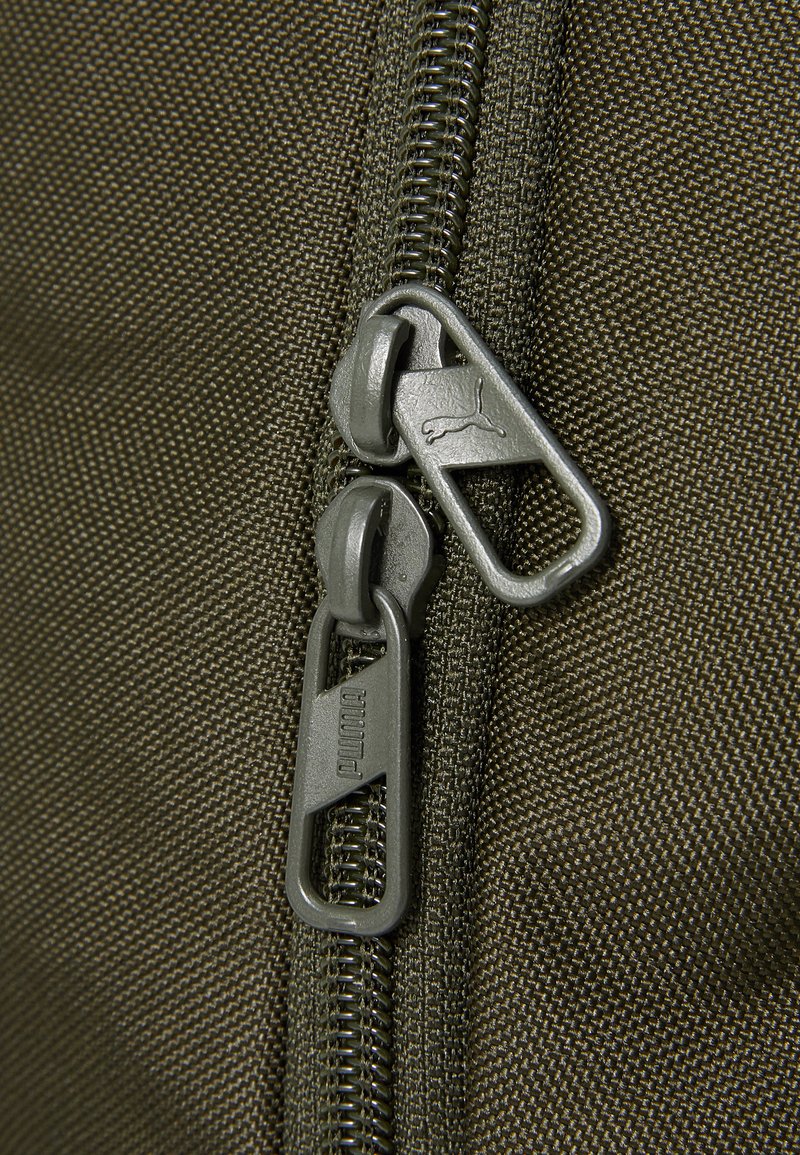 Sac en tissu vert olive avec deux fermetures éclair métalliques arborant le logo Puma, mettant en avant une surface texturée et des coutures durables.