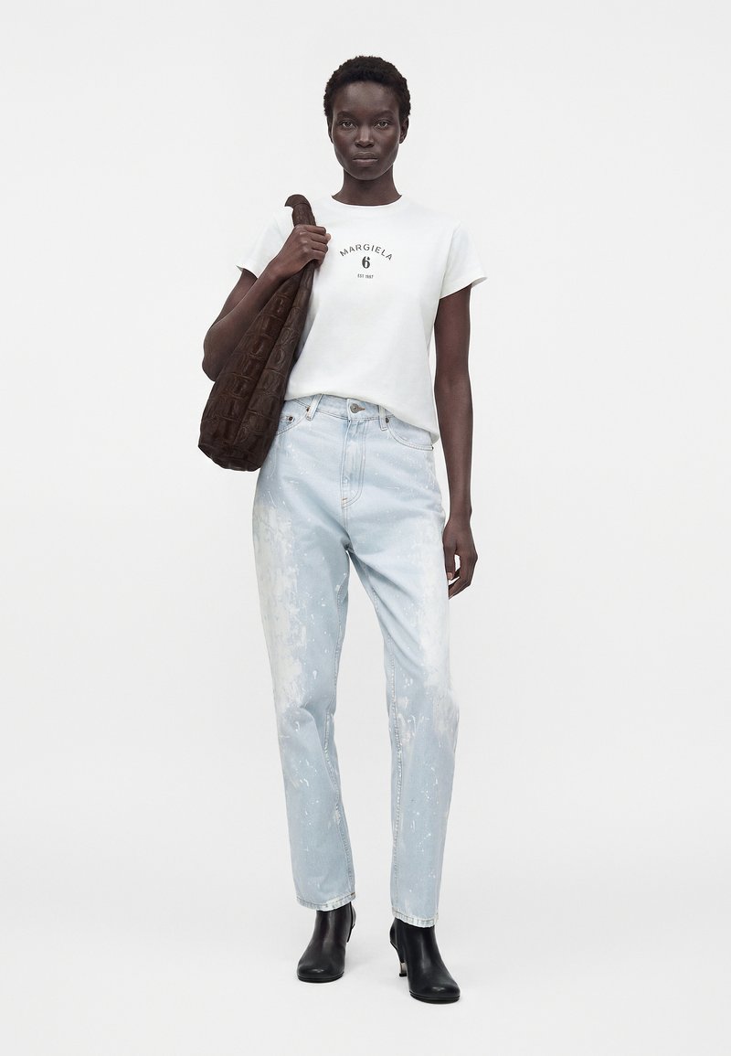 Woman wearing white logo t-shirt, light blue paint-splattered jeans, black ankle boots, holding large dark brown textured shoulder bag.