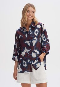 Kragenhemd in Maroon mit blau-weißem abstraktem Muster. Lange Ärmel hochgerollt, aus leichtem Stoff. Einfaches Kragendesign.