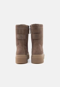 Brązowe zamszowe botki za kostkę z żebrowanymi gumowymi podeszwami, tylnym zamkiem błyskawicznym i przeszytymi pętelkami z logo "M OP", zaprezentowane na białym tle.