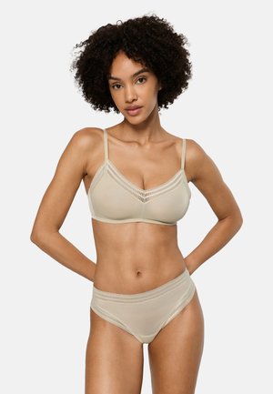 Vrouw met krullend haar, gekleed in een beige beha met kanten randen en bijpassend hoog tailleondergoed, staand met handen achter de rug tegen een witte achtergrond.