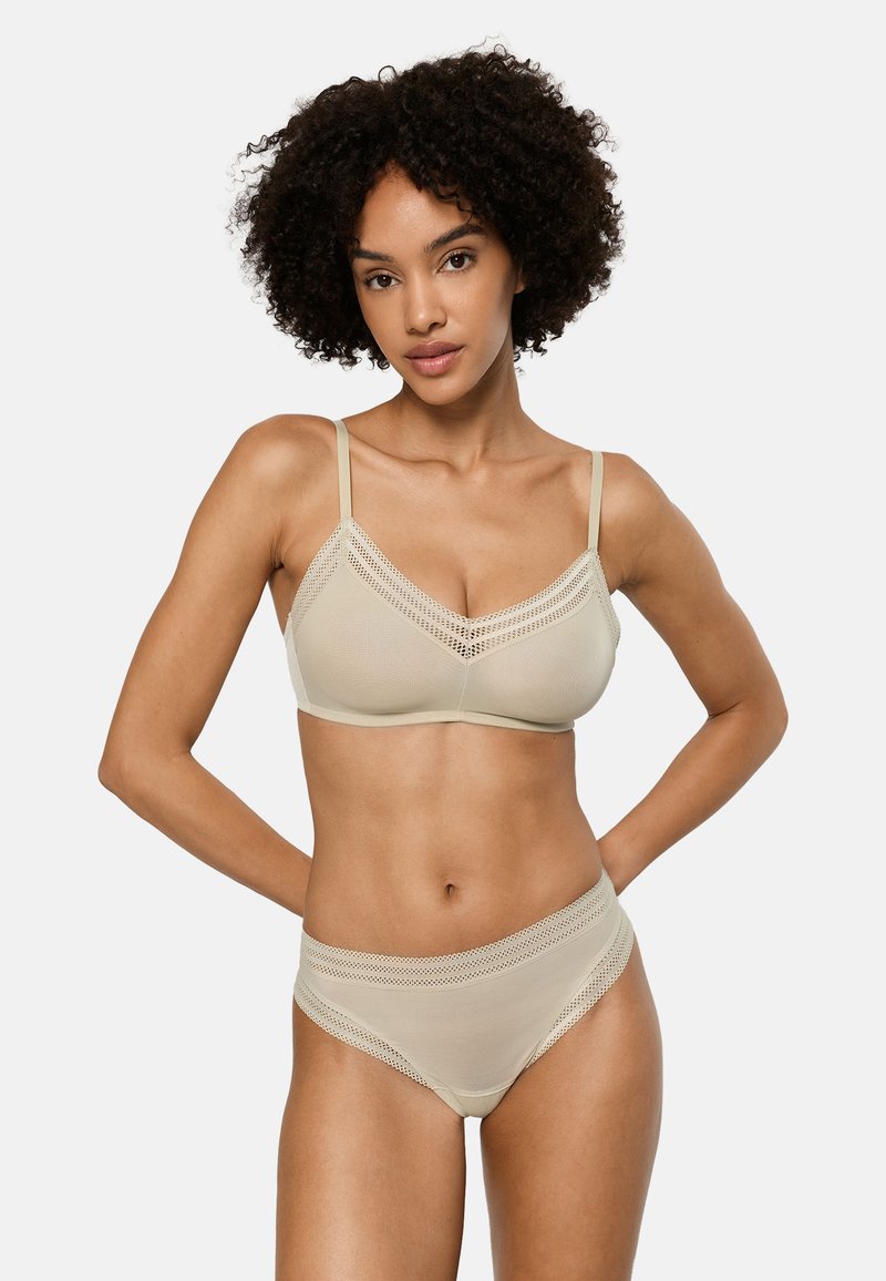 Frau mit lockigem Haar, die einen beigefarbenen BH mit Spitzenbesatz und passende High-Waist-Unterwäsche trägt, steht mit den Händen hinter dem Rücken vor weißem Hintergrund.