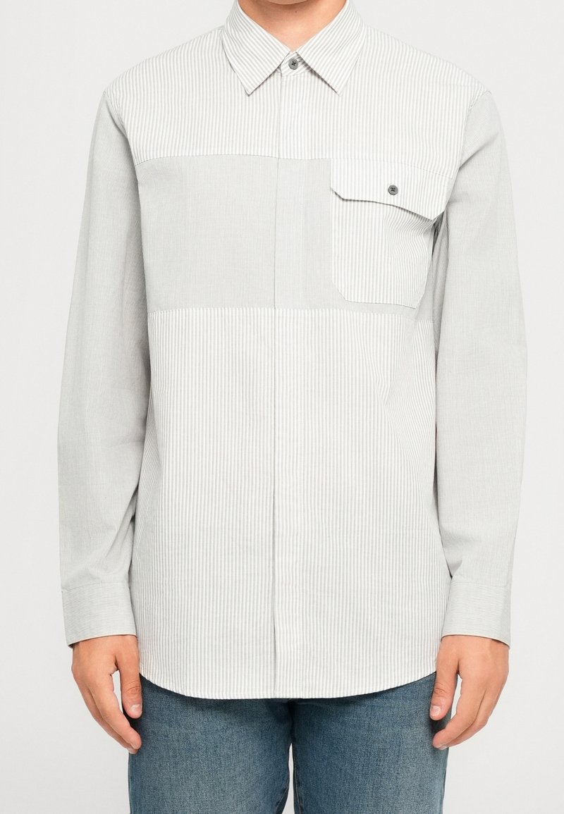 Homme portant une chemise à manches longues rayée gris clair et blanc, avec une poche poitrine à bouton unique, et un jean bleu.