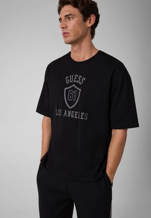 Jeune homme portant un t-shirt noir avec un texte gris "GUESS 81 LOS ANGELES" et un logo bouclier, debout les mains dans les poches, de profil.