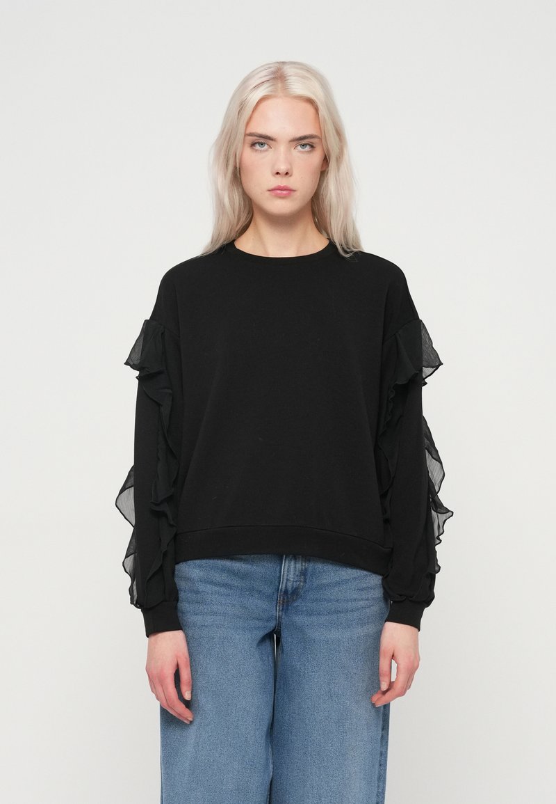 Czarny sweter z falbankowymi przezroczystymi rękawami, okrągłym dekoltem i luźnym krojem. W zestawie z jasnoniebieskimi spodniami o szerokich nogawkach.