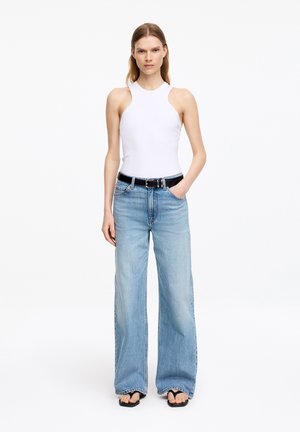 Kobieta ubrana w biały top bez rękawów, jasnoniebieskie jeansy o szerokich nogawkach z czarnym paskiem oraz czarne klapki typu japonki, stojąca z jedną ręką w kieszeni.