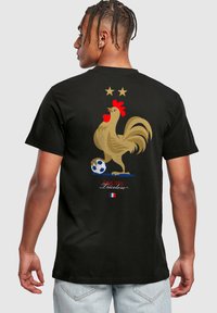 Sort bomulds t-shirt med et guld høne grafisk motiv, røde detaljer, to stjerner ovenfor og en fodbold nedenunder, med teksten "ÉQUIPE Tricolore".