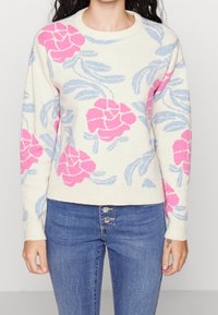 Strickpullover in Creme mit pinkem Blumenmuster und blauen Blättern. Verfügt über einen runden Ausschnitt und gerippte Bündchen. Kombiniert mit blauen Jeans.
