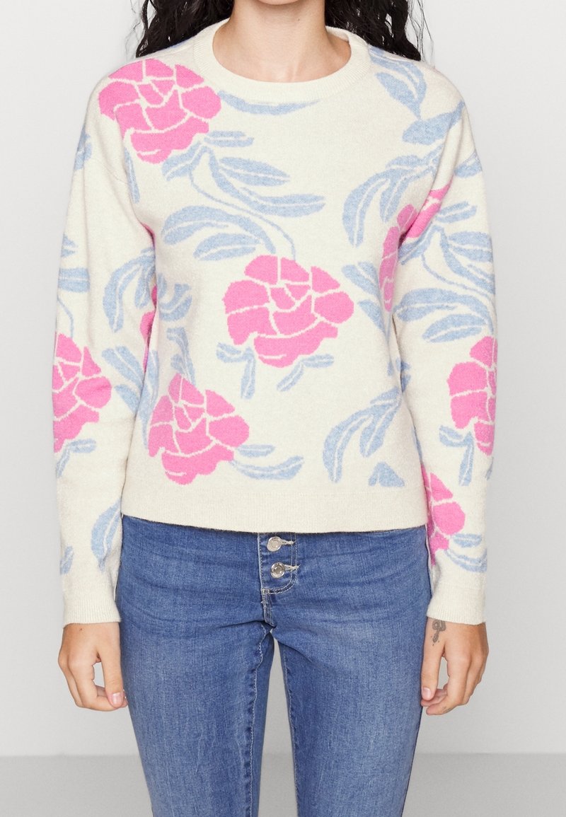 Strickpullover in Creme mit pinkem Blumenmuster und blauen Blättern. Verfügt über einen runden Ausschnitt und gerippte Bündchen. Kombiniert mit blauen Jeans.