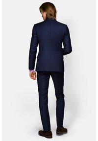 Granatowy, dopasowany garnitur o fakturowanym wykończeniu, z jednym rozcięciem, spodniami slim-fit i ciemnobrązowymi butami. Akcenty obejmują jasnoróżowy mankiet koszuli.