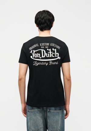 Oseba s kratkimi črnimi lasmi, oblečena v črno majico Von Dutch z belim napisom "Original Kustom Kulture 1929-1992 Legendary Brand" na hrbtu.