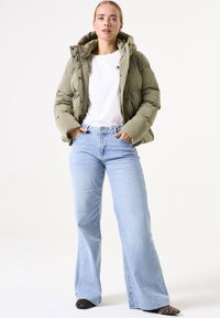 Chaqueta acolchada verde oliva con capucha, camiseta blanca, vaqueros acampanados de un azul claro y zapatos con estampado animal. Diseño simple y acolchado con cierres de botón.