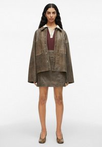 Modelka stoi w neutralnym otoczeniu, ubrana w brązową, oversize'ową koszulę, bordowy polo oraz dopasowaną spódnicę, do których ma na sobie brązowe szpilki.