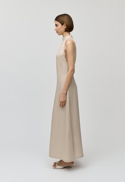 Beiges ärmelloses Kleid mit hohem Kragen, tailliertem Oberteil und fließendem Saum. Glatte Textur mit seitlichen Ausschnitten. Modell trägt neutrale, hohe Schuhe.