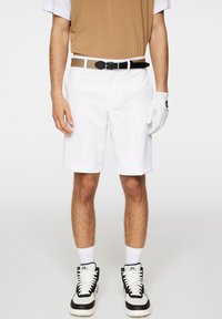 Białe spodenki golfowe o gładkiej fakturze, z przodu z plisami i czarnym paskiem. W zestawie z brązowo-białą koszulą i czarnymi sneakersami.