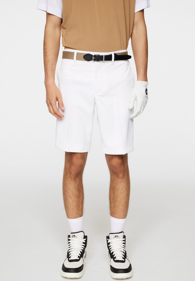 Białe spodenki golfowe o gładkiej fakturze, z przodu z plisami i czarnym paskiem. W zestawie z brązowo-białą koszulą i czarnymi sneakersami.