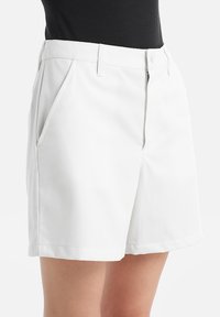 Pantaloni corti bianchi realizzati in tessuto liscio, con pieghe frontali, tasche laterali e orlo pulito. Nessun abbellimento o motivo visibile.