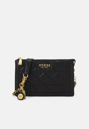 Pochette noire matelassée Guess avec fermeture éclair dorée, logo et breloques décoratives en chaîne sur fond blanc.