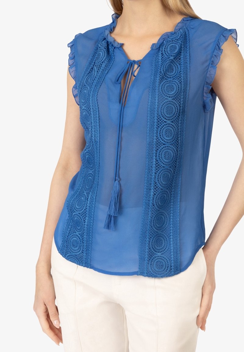 Femme portant un chemisier bleu transparent sans manches avec des empiècements en dentelle, des épaules volantées et un lien à pompons au col, associé à un pantalon blanc.