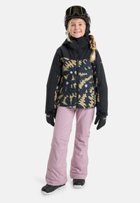 Skioutfit mit einer schwarzen Jacke im Camouflage-Muster in Gold und Lila, kombiniert mit hellrosa Hose und schwarzen Schneestiefeln.