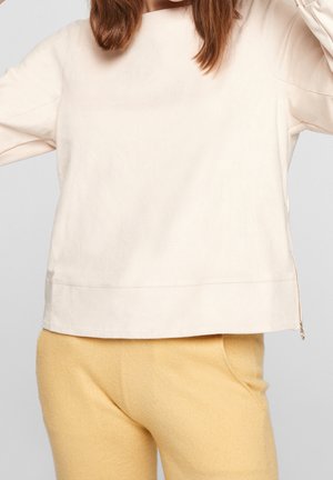 Femme portant un haut ample crème à manches longues avec une fermeture éclair latérale et un pantalon en polaire jaune doux sur un fond uni.