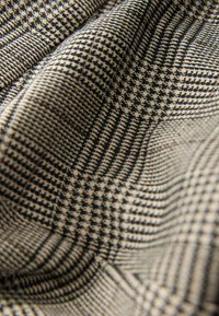 Nærbillede af beige og sort stof foldet, med et detaljeret houndstooth mønster i varierende størrelser og komplekse teksturer.