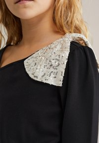 Crna majica s teksturiranim, šarenim detaljem na ramenu u krem i srebrnim tonovima, sa okruglim izrezom i mekanom tkaninom.