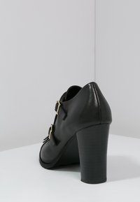 Zapato negro de tacón con acabado brillante, que presenta dos hebillas doradas y un tacón robusto. Textura suave con un diseño de punta afilada.