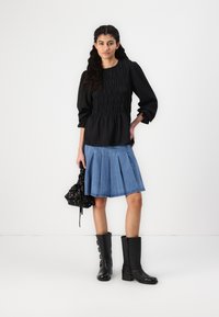 Black smocked blouse with three-quarter sleeves, paired with a pleated denim skirt. Black ankle boots and a black patterned drawstring bag complete the look.