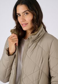 Beige gevoerde jas met een ruitvormig doorgestikt patroon, hoge kraag met zachte bruine bontafwerking, ritssluiting aan de voorkant en een slank silhouet.