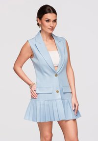 Femme portant une robe blazer sans manches bleu clair avec des boutons dorés, une jupe plissée, un bandeau blanc et des bracelets dorés sur un fond uni.
