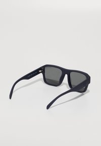 Occhiali da sole blu navy con finitura opaca. Forma quadrata con lenti grigie e sottili bracci curvi. Branding discreto sul lato.