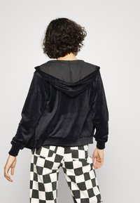 Personne avec des cheveux bouclés portant un sweat à capuche en velours noir et un pantalon à carreaux noir et blanc, vu de dos contre un fond uni.