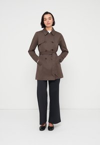 Καφέ διπλής όψης παλτό με ζώνη, με γιακά και δύο εμπρόσθιες τσέπες, συνδυασμένο με μαύρο παντελόνι με ρίγες και μαύρα παπούτσια.