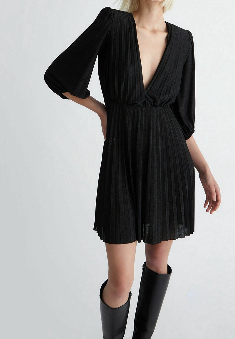 Robe noire plissée avec un décolleté en V profond et des manches ballon. Tissu doux et texturé avec une taille cintrée, un ourlet au-dessus du genou et une silhouette fluide.