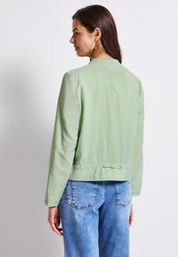 Světle zelená bomber bunda s texturovaným povrchem, s žebrovaným límcem a detaily vázání na zadní straně, doplněná světle modrými džínami.