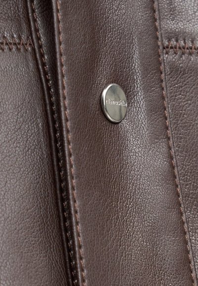 Közelkép barna bőrről, varrással és egy kis kerek fémbotton, amelyen az "Nanushka" felirat szerepel.