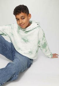 Chłopiec z krótkimi, kręconymi włosami, ubrany w zieloną bluzę w tie-dye i niebieskie dżinsy, siedzący na białej podłodze z zrelaksowanym wyrazem twarzy.