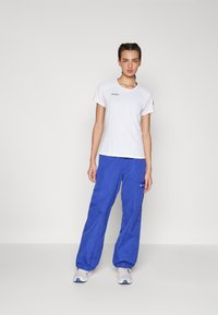 T-shirt branca de mangas curtas com um logótipo, combinada com calças cargo azuis de corte largo, apresentando bolsos e um detalhe de marca. Ténis completam o visual.