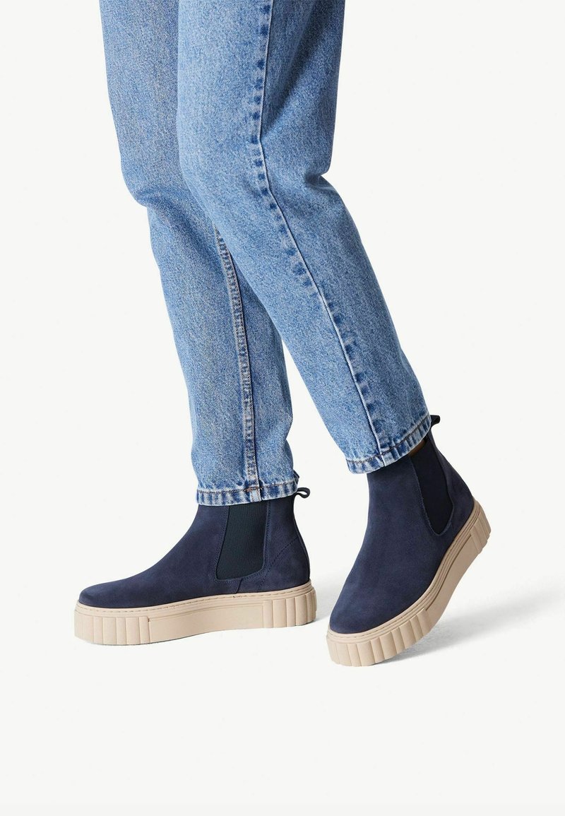 Personne portant un jean en denim bleu clair et des bottines Chelsea en daim bleu marine avec des semelles en caoutchouc beige épaisses.